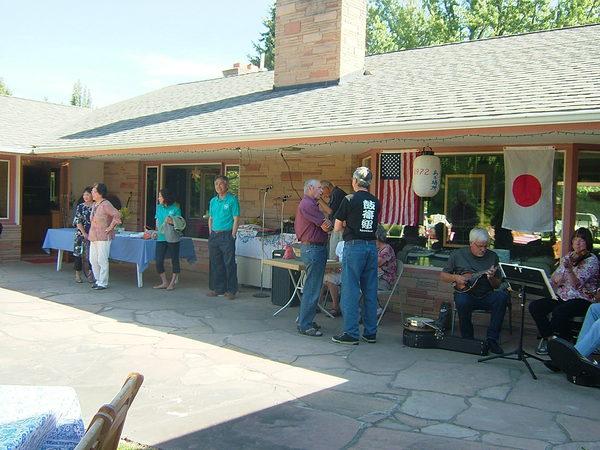 日本とアメリカの国旗が飾られた店の前で演奏をしている人の隣で男性2人がお話ししていてその横にはマイクが2本用意され、4人の人がたっている写真